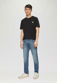 Schwarzes Baumwoll-T-Shirt mit gelbem Logo, blauwe Slim-Fit-Jeans und weißen Sneakern mit grauen Akzenten und beigen Sohlen, vor einem hellen Hintergrund stehend.