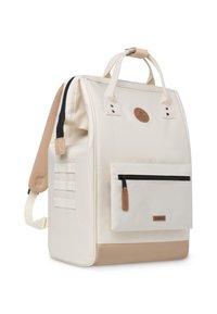 Mochila de tela crema con detalles en tonos marrones, forma rectangular, asa superior, tirantes ajustables, bolsillo frontal con cremallera y detalle de logo.
