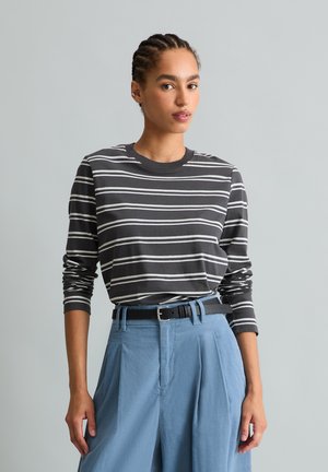 Femme aux cheveux tressés portant une chemise à manches longues rayée noir et blanc rentrée dans un pantalon bleu clair taille haute avec une ceinture noire.