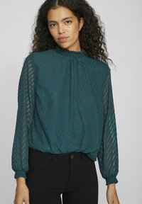 Blusa strutturata color teal con un alto colletto arricciato, maniche lunghe trasparenti e polsini elasticizzati; presenta un motivo a zigzag diagonale e un orlo arricciato.