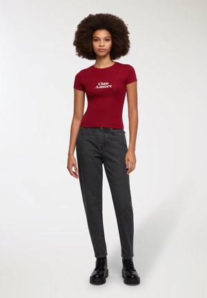 T-shirt bordeaux con testo "Ciao Amore", abbinata a jeans neri a vita alta e stivali neri con lacci, su uno sfondo neutro.