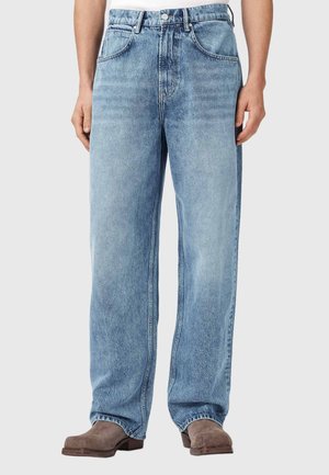 Lyseblå løstsiddende jeans båret med brune ruskindsstøvler, vist fra talje til fødder på en stående person mod en ensfarvet baggrund.