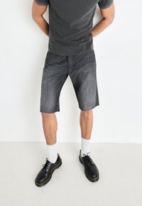 Shorts de mezclilla negra con dobladillo deshilachado, combinados con calcetines blancos y zapatos negros con un acento de costura amarilla. Camiseta gris ajustada.