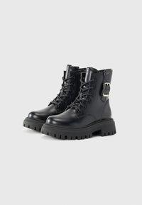 Botas de tobillo de cuero negro con un diseño de cordones, acento de hebilla de metal y suela de goma gruesa con surcos profundos.