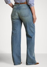 Sieviete, kas valkā augstās jostasvietas taisnu kāju zilas džinsas, brūnu jostu, svītrainu kreklu ar uzlocītām piedurknēm un brūnas ar punktu norādītu apavus, stāvot ar muguru pret skatītāju.