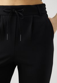 Svarte joggebukser laget av mykt stoff, med snøring i livet, folder og sidelommer. Glatt tekstur med minimalistiske detaljer.