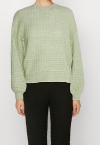 Ljust grönt, strukturerat tröja med rund hals, ribbade ärmar och nederkant, tillsammans med svarta, strukturerade byxor. Materialen verkar mjuka och stickade.