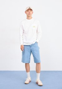 Homme portant une casquette beige, une chemise blanche à manches longues, un short en denim bleu clair, des chaussettes blanches et des baskets blanches avec des rayures roses, se tenant sur un sol bleu.