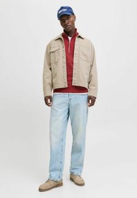 Giacca beige con bottoni frontali, maglione rosso con zip sotto, jeans azzurri chiari e scarpe marrone chiaro. Berretto blu con testo bianco sopra.