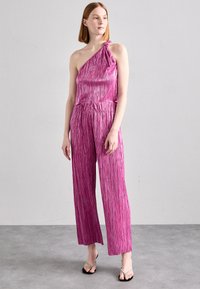 Combinaison à une épaule en rose vif avec des plis verticaux, taille élastique et coupe large. Présente un détail noué à l'épaule.
