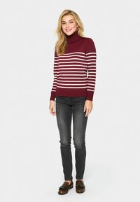 Pull à col roulé bordeaux avec des rayures blanches, poignets et ourlet côtelés. Porté avec un jean foncé et des mocassins marron, le modèle est debout sur un fond neutre.