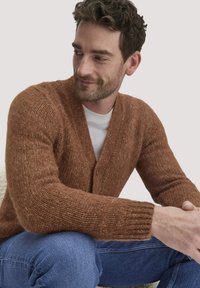 Brauner, strukturierten Cardigan mit V-Ausschnitt und Knopfdetails. Strickstoff, gerippte Bündchen. Wird über einem weißen T-Shirt getragen, kombiniert mit blauen Jeans.