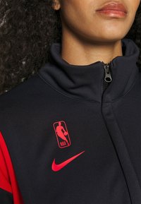 Personne portant un haut zippé noir et rouge avec un logo NBA rouge et un swoosh Nike sur la poitrine, montrant le cou et le bas du visage.