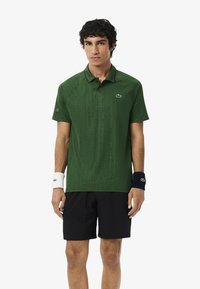 Uomo che indossa una polo verde scuro, pantaloni corti neri e braccialetti non abbinati, in piedi su uno sfondo bianco uniforme.
