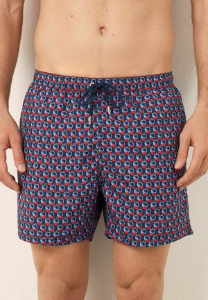 Mann in marineblauen Badeshorts mit rot und hellblauem geometrischen Fischmuster und einem marineblauen Zugbandgürtel, der an einer Wand steht.
