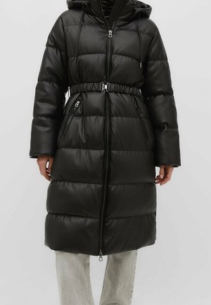 Czarna pikowana kurtka puchowa z kapturem, paskiem w talii, kieszeniami na zamek i przednim zamkiem noszona na jasnych spodniach.