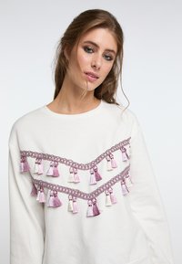 Felpa bianca con un design a zigzag di rifiniture intrecciate viola e con frange in diverse tonalità di viola e bianco sul davanti.