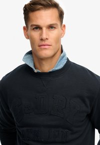 Mørkeblå sweatshirt med præget skrift "S&D DRY" og "ATH.JPN." Lagdelt over en lyseblå skjorte med krave. Blødt stof, afslappet design.