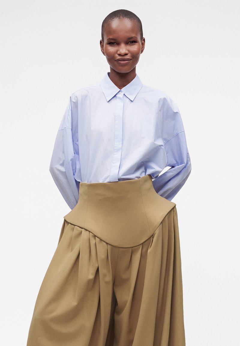 Personne souriante aux cheveux courts portant une chemise bleu clair boutonnée rentrée dans un pantalon beige large plissé taille haute, sur un fond uni.