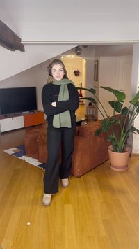 Maglione nero, sciarpa verde a coste e pantaloni neri a gamba larga. Scarpe di colore chiaro. Ambiente accogliente con una pianta e un divano di velluto a coste marrone.