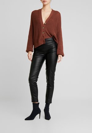 Kvinde iført en brun strikket knapcardigan, sorte bukser i imiteret læder og sorte ankelstøvler med høje hæle stående på en grå baggrund.