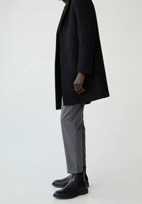 Schwarzer langer Mantel, graue maßgeschneiderte Hose und schwarze Stiefeletten. Der Mantel hat einen geraden Schnitt; die Hose ist maßgeschneidert und hat einen Saum.