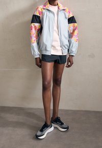 Chaqueta atlética de color gris claro con mangas de teñido anudado multicolor, detalles en negro, combinada con una camiseta rosa y pantalones cortos oscuros, llevada con zapatillas negras.