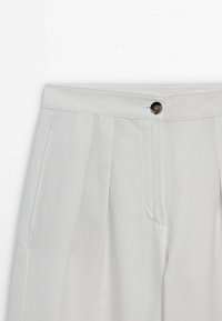 Pantalon gris clair taille haute avec plis à l'avant, poches latérales, fermeture bouton et ceinture.