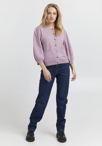 Lila stickad cardigan med puffärmar, framfickor och svarta knappar, tillsammans med mörka jeansbyxor och svarta skor.