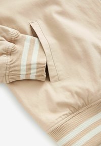 Felpa beige con finitura testurizzata, caratterizzata da un polsino a costine accentuato da righe bianche e da una tasca laterale per praticità.
