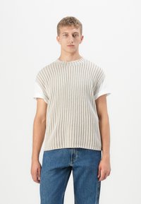 Haut tricoté côtelé beige clair à manches courtes, superposé à un T-shirt blanc. Porté avec un jean en denim bleu. Design simple et texturé.