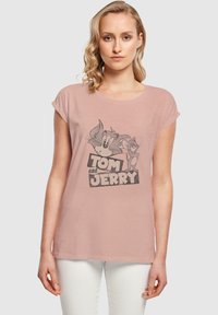 Heled roosa T-särk rullitud varrukatega, millel on halltooni illustration Tomist ja Jerryst, ning julgelt kirjutatud "TOM JA JERRY" tekst all.