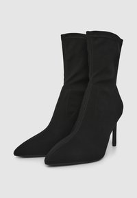 Czarne botki za kostkę z gładką zamszową fakturą, spiczastym noskiem i wysokim obcasem w kształcie szpilki. Posiadają dekoracyjne przeszycia i dopasowany krój.