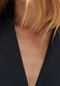 Černá tkanina s designem ve tvaru V, hladká textura a viditelné tóny pleti s jemnými pihami. Vlasy volně vlnité.