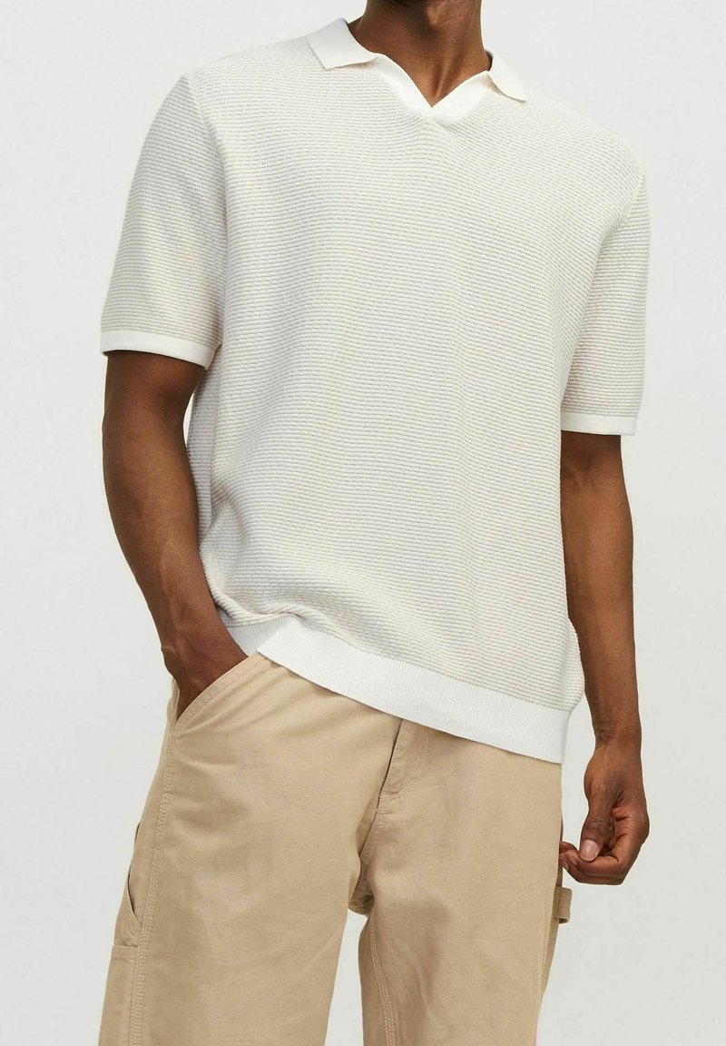 Pletený polo s krátkým rukávem v světle béžové barvě s texturovaným vzorem, s výstřihem ve tvaru V a bílými ribbovanými límečkem a manžetami. Volnější střih.