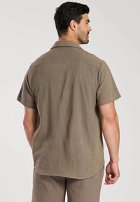 Chemise marron à manches courtes en tissu texturé. Présente un motif de slub subtil et un col classique, avec un ourlet arrondi à l'arrière.