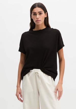 Woman with straight dark hair wearing a loose black short-sleeve top and light beige drawstring pants against a plain light background.