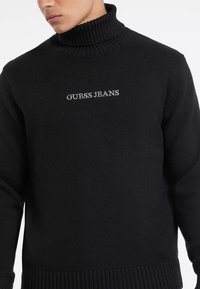 Maglione nero a collo alto in maglia con polsini e orlo a costine, con "GUESS JEANS" ricamato in bianco sul petto.