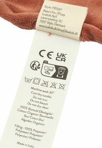 Brązkowa etykieta materiałowa z instrukcjami pielęgnacji, oznaczeniami CE i UKCA. Materiał: 100% bawełna organiczna, wypełnienie: 100% poliester. Wyprodukowano w Indiach.