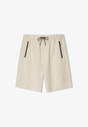 Beige Shorts in Knielänge mit elastischem Bund, schwarzem Kordelzug und schwarzen Reißverschlusstaschen an den Seiten vor weißem Hintergrund.