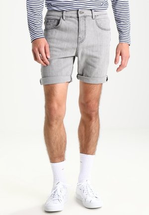 Homme portant un short en jean gris clair retroussé, des baskets blanches, des chaussettes blanches et une chemise à manches longues rayée bleu marine et blanc.