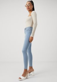 T-shirt de manga longa em tom beige com textura canelada, combinado com jeans ajustados azuis claros e sandálias de salto bloco brancas. A modelo posa de lado contra um fundo claro.