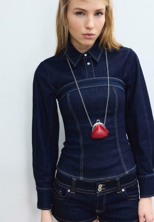 Ung kvinde iført mørkt denimoutfit med langærmet top og shorts, accessoriseret med en lille rød pung på en sølvkæde.