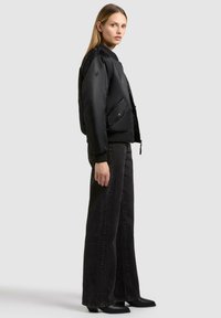 Schwarze Bomberjacke mit Reißverschluss und vorderen Taschen, kombiniert mit hoch sitzenden, schwarzen weitem Jeans und schwarzen Stiefeletten mit Absatz.