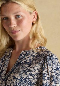 Tumes lillemustriga pluus valgete botaaniliste printidega, ümmargune kaelus ja kogutud puhvvarrukad. Kullakarva rõngaste kõrvarõngad täiustavad mudeli välimust.