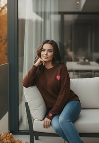 Brązowy sweter z różowym sercem, w połączeniu z niebieskimi dżinsami. W tle znajdują się jasnokryte kanapa i duże szklane okna.