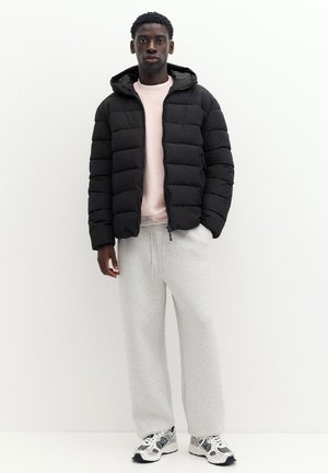 Young man standing wearing a black puffer jacket, light pink sweatshirt, gray sweatpants, and white sneakers against a plain white background.