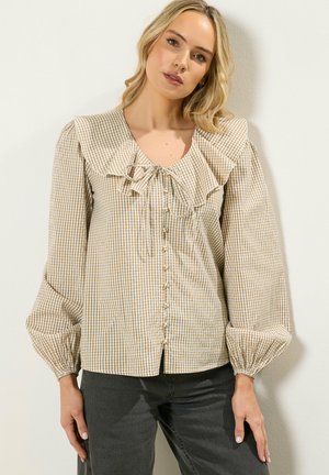 Lyshåret kvinde iført beige gingham-bluse med stor flæsekant, knapper foran og lange puffede ærmer, kombineret med mørkegrå bukser.