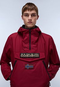 Jonge man met kort haar die een rode Napapijri anorakjas draagt met zwarte ritsen en een Noorwegen-vlagpatch, staande tegen een effen achtergrond.