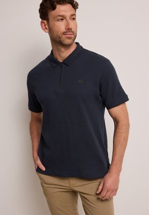Homme portant un polo bleu marine à manches courtes avec un col à fermeture éclair et un pantalon beige, debout avec une main dans la poche, devant un fond uni.